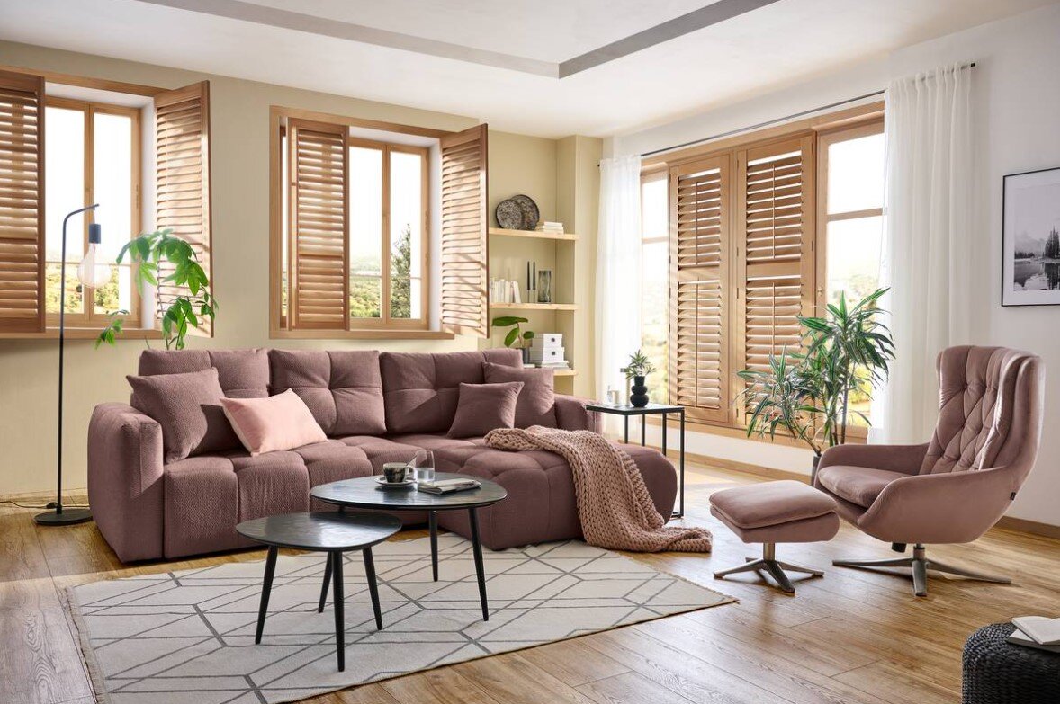 Hom´in Hoekbank London Een gezellig interieur van een woonkamer met een grote, roze hoekbank, die vanaf de zijkant wordt getoond. Op de voorgrond staan twee ronde salontafels op een geometrisch tapijt. Rechts in beeld staat een comfortabele fauteuil met een bijpassend voetenban