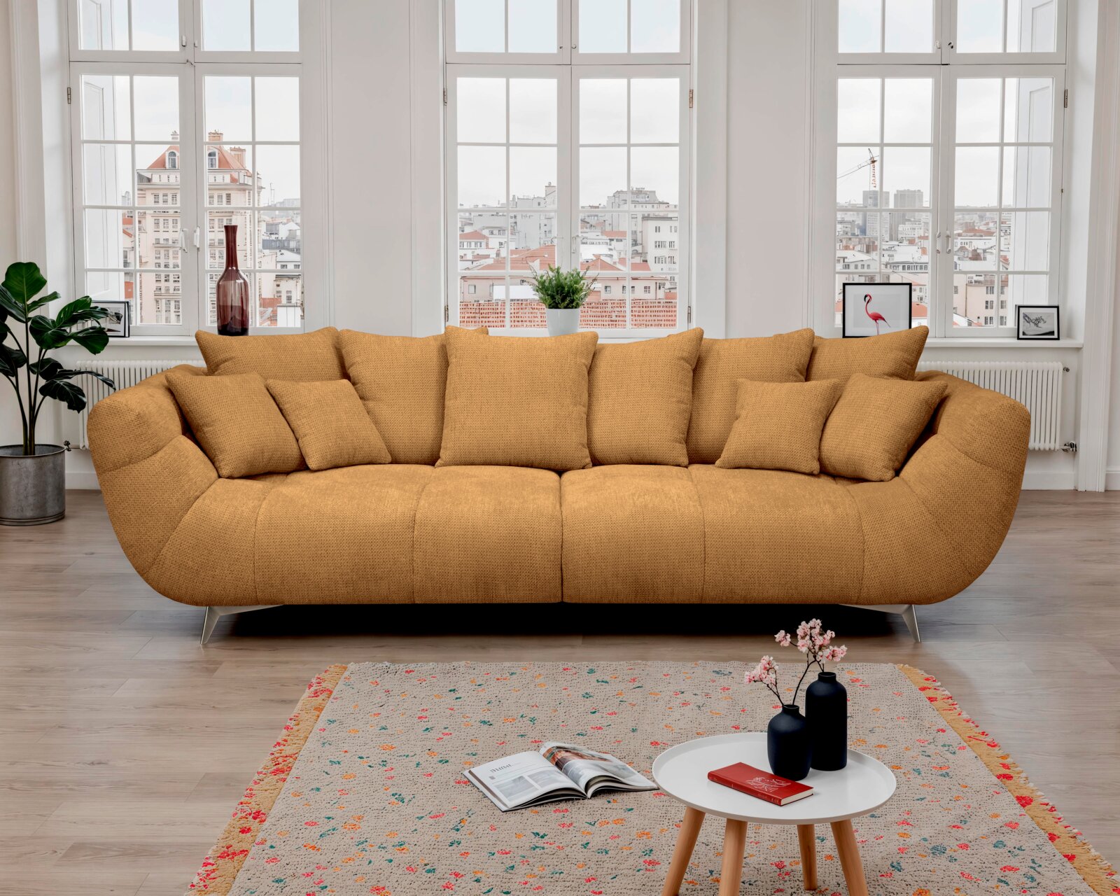 Modernes, ockerfarbenes 3-Sitzer-Stoffsofa mit vielen Kissen und Metallfüßen in hellem Wohnzimmer vor großen Fenstern, Frontansicht auf Augenhöhe
