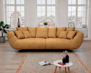 Modernes, ockerfarbenes 3-Sitzer-Stoffsofa mit vielen Kissen und Metallfüßen in hellem Wohnzimmer vor großen Fenstern, Frontansicht auf Augenhöhe