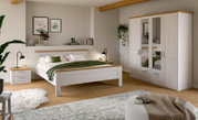 Modernes Schlafzimmer-Set in Weiß mit Holz-Akzenten: Doppelbett mit Nachttisch und Kleiderschrank mit Spiegeltüren, fotografiert in schräger Frontansicht aus dem Raum.