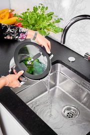 Topf mit Glasdeckel wird in der Küche über einem Waschbecken aus einer seitlichen Perspektive abgegossen, frisches Gemüse im Topf sichtbar.