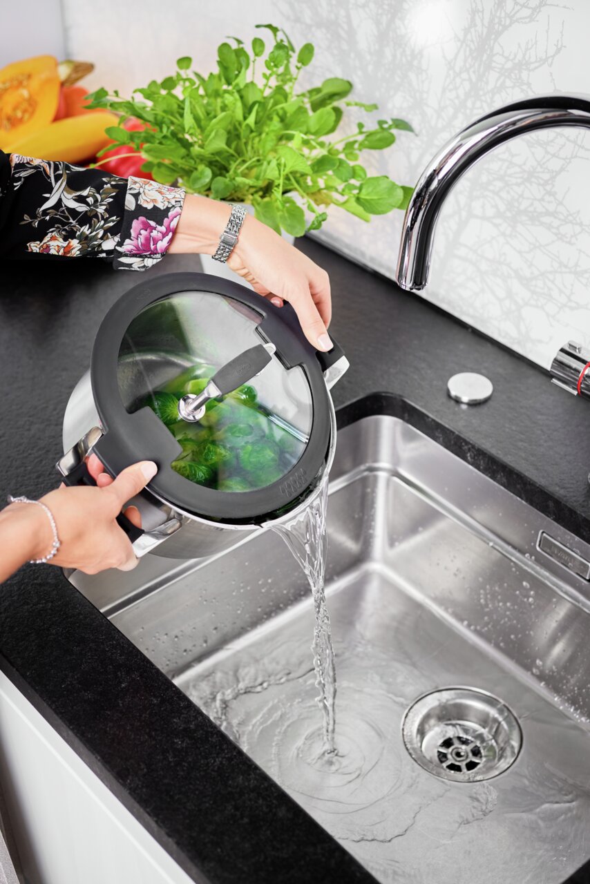 Topf mit Glasdeckel wird in der Küche über einem Waschbecken aus einer seitlichen Perspektive abgegossen, frisches Gemüse im Topf sichtbar.