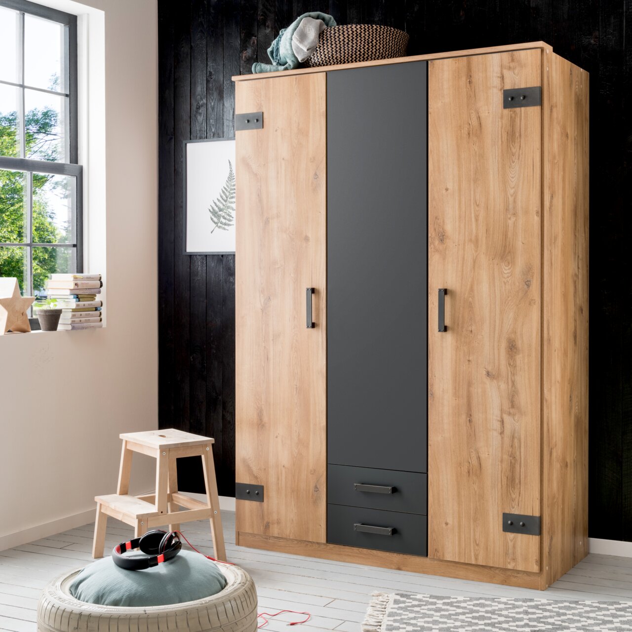 Drehtürenschrank mit drei Türen in Holzoptik und grauem Mittelteil, frontal fotografiert in einem modernen Raum mit Fenster und Dekoration.