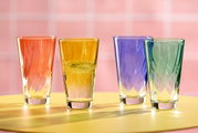 Vier bunte Trinkgläser in den Farben Rot, Gelb, Blau und Grün auf einem gelben Tisch vor einem rosa Hintergrund, seitliche Perspektive.