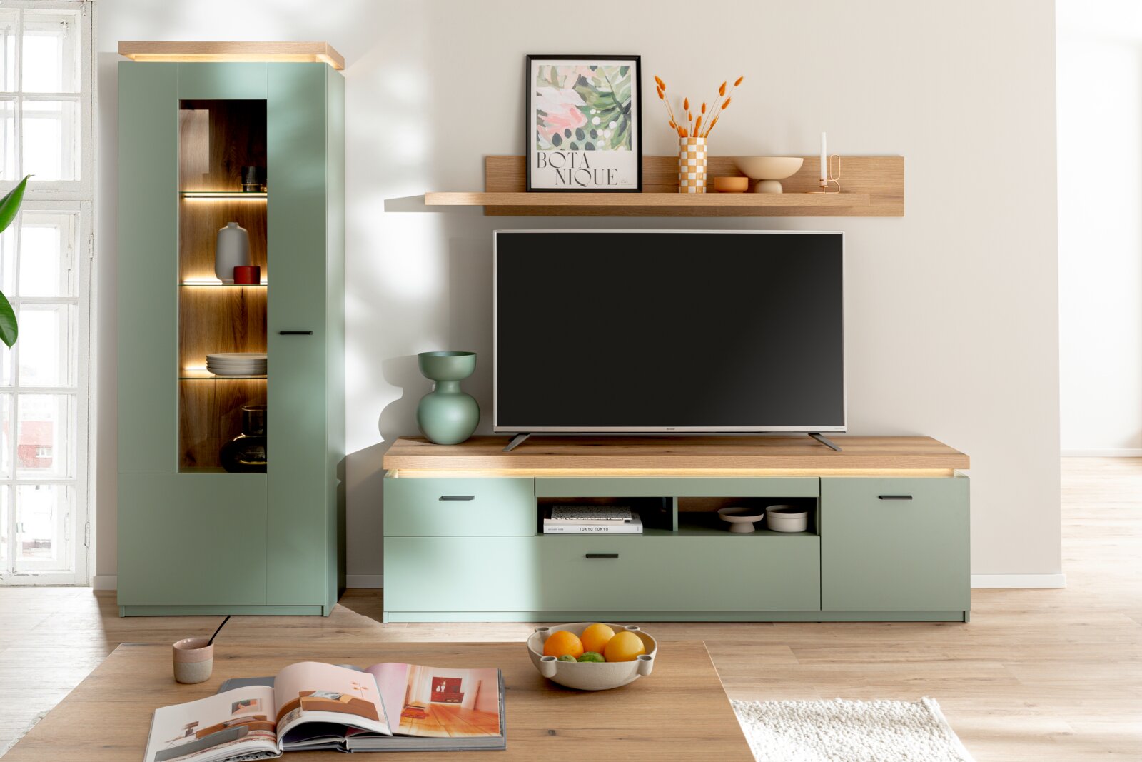 Z2 Vitrine MONITZ Frontale Perspektive auf moderne Wohnwand in Salbeigrün mit Holzoptik, TV-Lowboard und hohem Vitrinenschrank mit LED-Beleuchtung sowie Wandboard über einem Fernseher im hellen Wohnzimmer.
