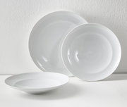 Drei weiße, glänzende Porzellanteller (zwei große, ein tiefer) vor heller Wand, fotografiert in leichter Schrägansicht von vorne.