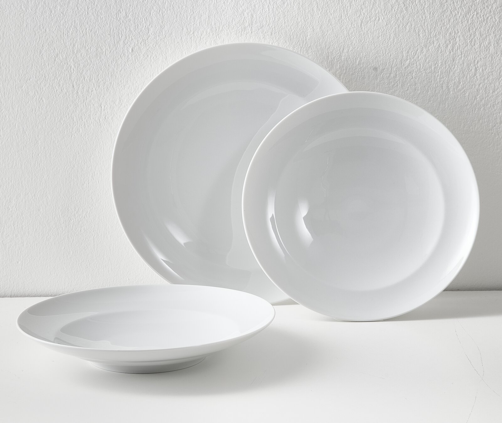 Drei weiße, glänzende Porzellanteller (zwei große, ein tiefer) vor heller Wand, fotografiert in leichter Schrägansicht von vorne.