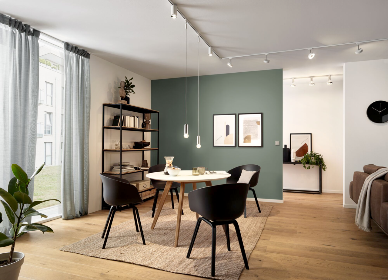 Moderne Wohnzimmerszene mit Schienensystem an der Decke, das mehrere verstellbare Leuchten trägt. Perspektive von der Seite, die den Raum mit Tisch, Stühlen und Regal zeigt.