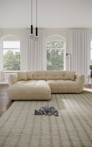Beige Ecksofa in einem hellen Wohnzimmer mit großen Fenstern, zwei Kissen und einer schlafenden Katze auf einem Teppich. Perspektive von vorne.
