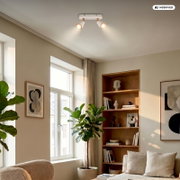 Moderne Deckenleuchte mit zwei verstellbaren Spotstrahlern in Weiß und Holzoptik, eingeschaltet, in einem hellen Wohnzimmer – Perspektive: Blick nach oben zur Decke aus Raumecke
