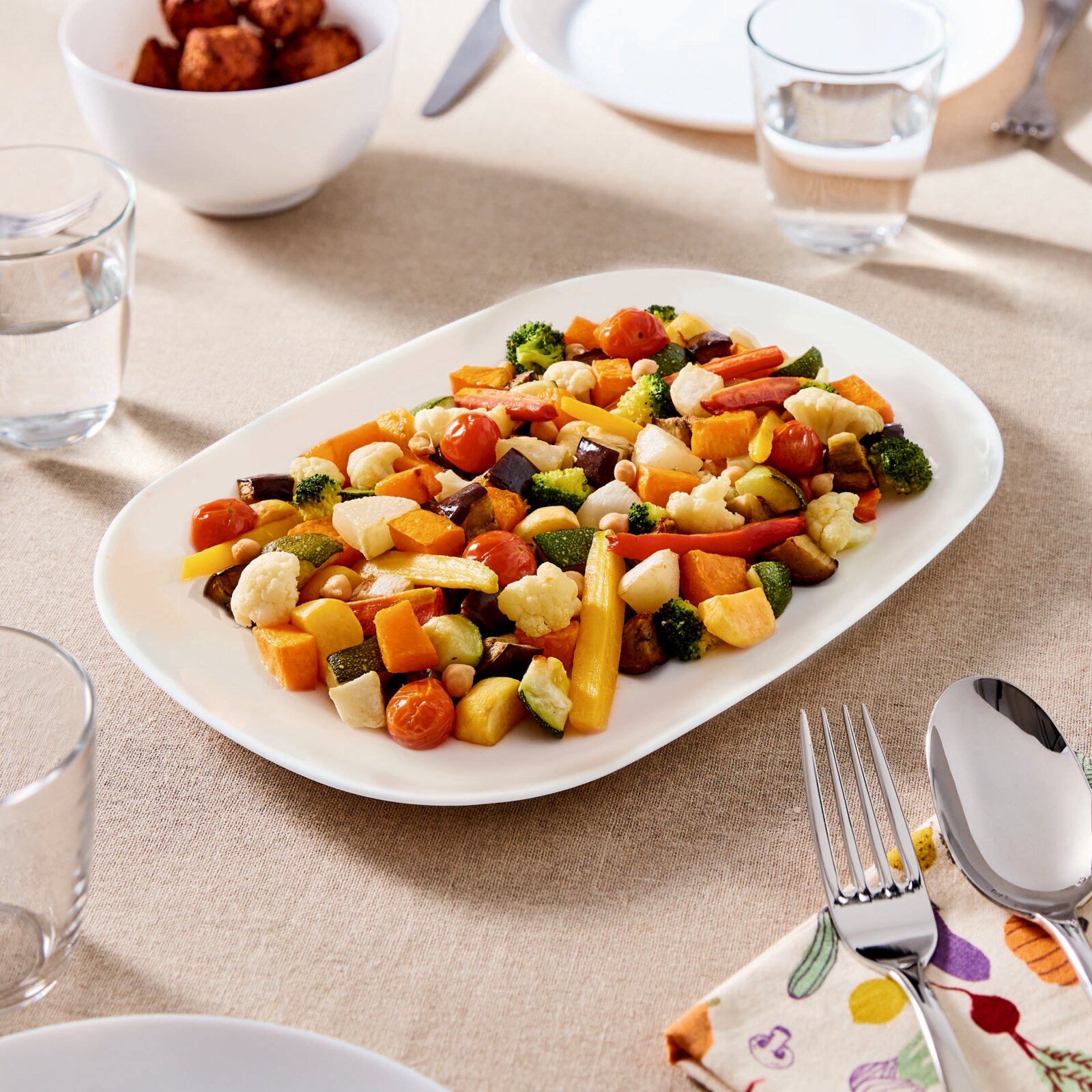 Bunte Gemüsemischung auf weißer ovaler Servierplatte (Brokkoli, Blumenkohl, Paprika, Zucchini, Aubergine, Tomaten) auf gedecktem Tisch, fotografiert aus leicht schräger Draufsicht.