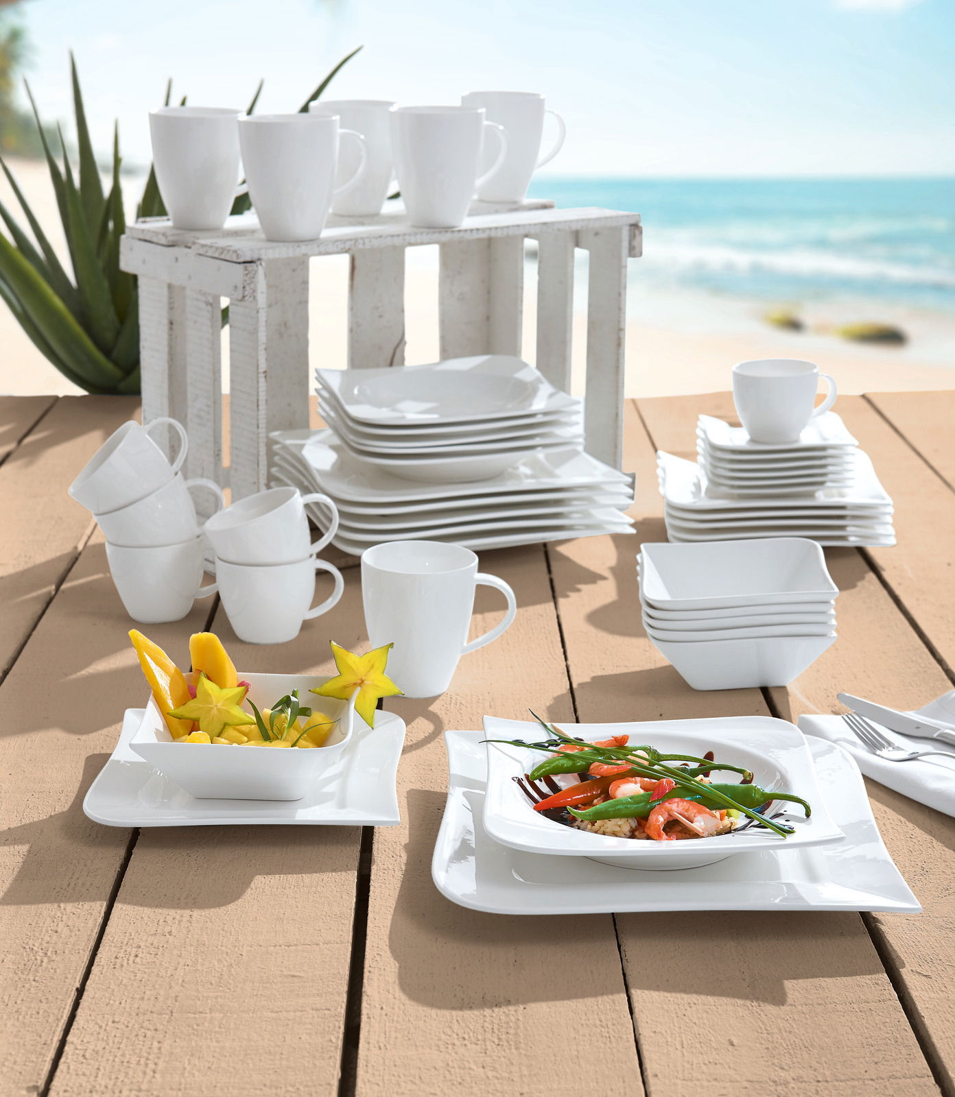 Kombiservice 42-teilig Melodie, bestehend aus weißen Tassen, Tellern und Schalen, auf einem Holztisch mit Strandhintergrund präsentiert. Perspektive: seitlich von oben.