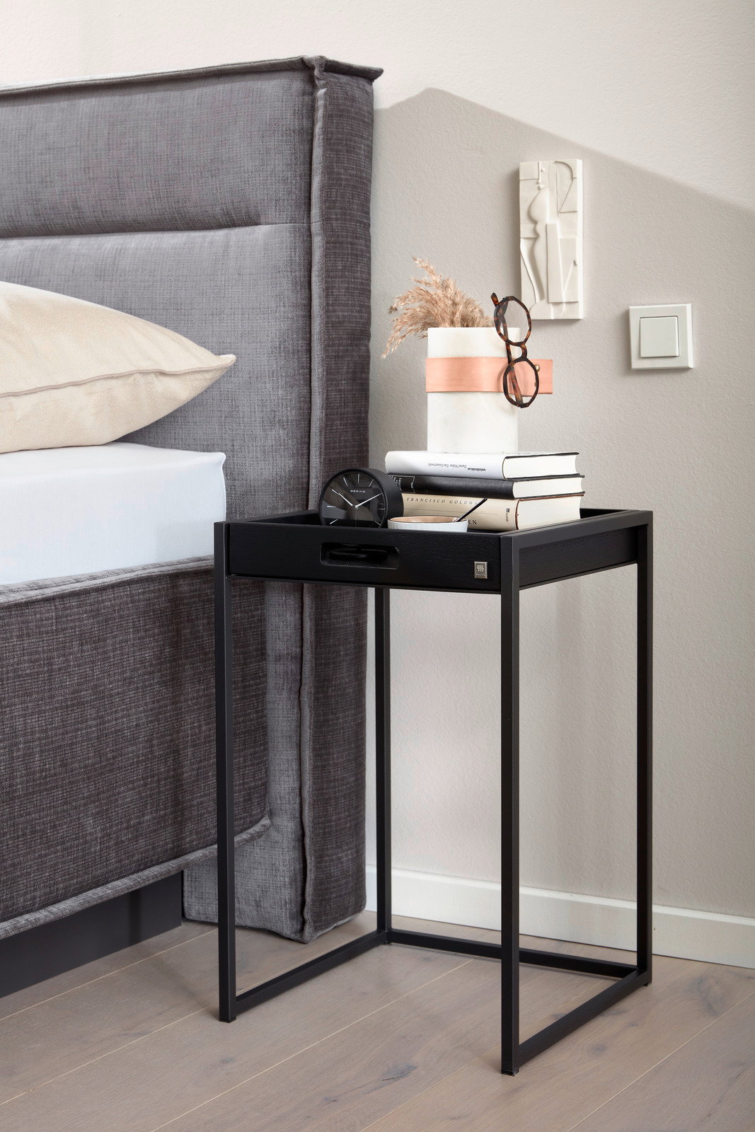 Moderner schwarzer Beistelltisch aus Metall neben einem Bett, dekoriert mit Büchern, einer Vase und einer Brille, seitliche Perspektive.
