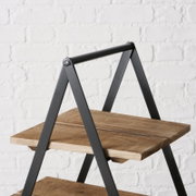 Nahaufnahme einer Etagere Valentina mit zwei Holzböden und schwarzem Metallrahmen vor einer weißen Ziegelwand.