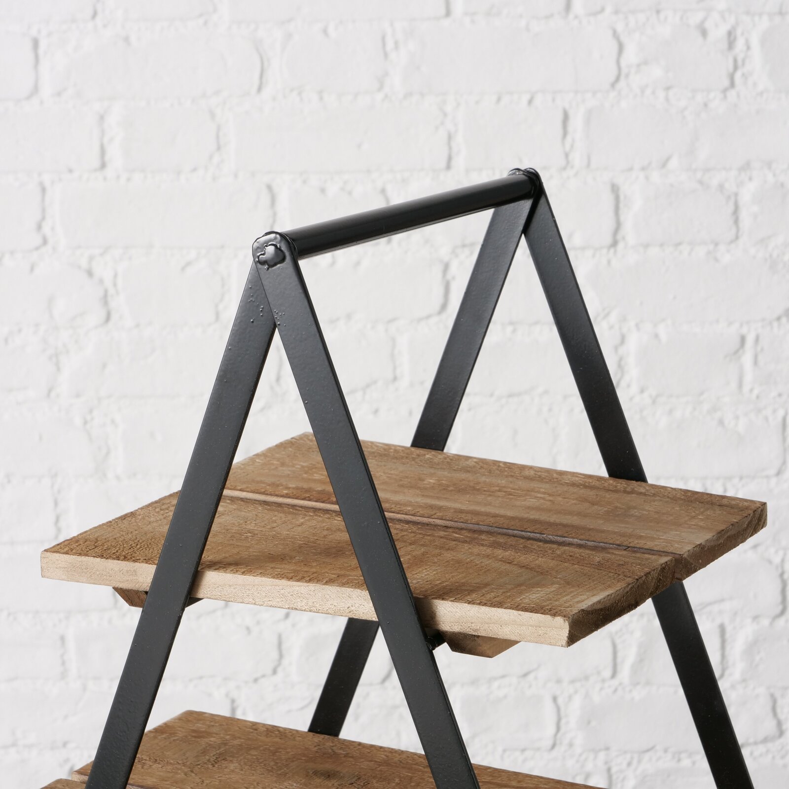Nahaufnahme einer Etagere Valentina mit zwei Holzböden und schwarzem Metallrahmen vor einer weißen Ziegelwand.