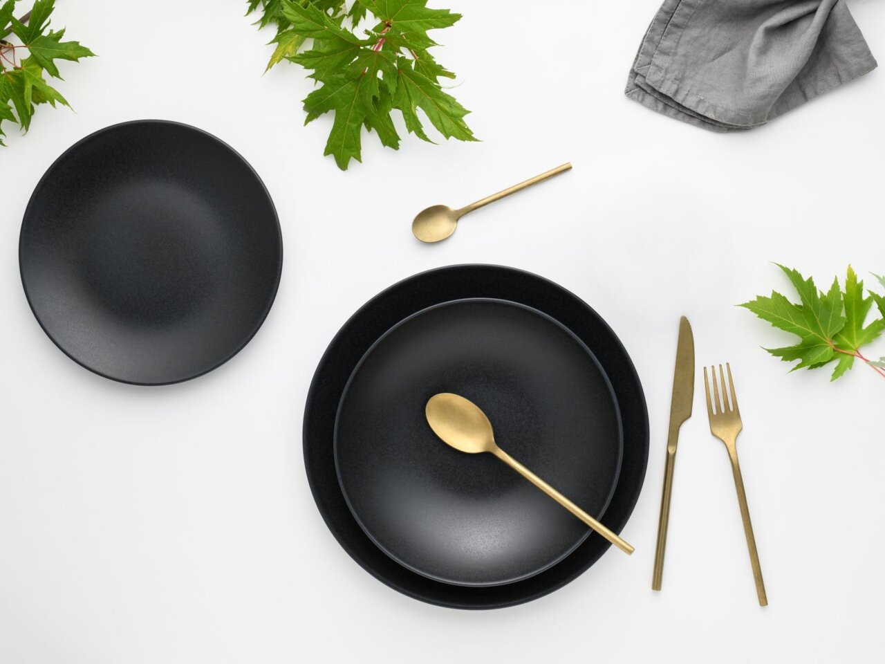 Zwart servies Soft Touch, 12-delig, met goudkleurig bestek, gefotografeerd vanuit vogelperspectief, versierd met groene bladeren.