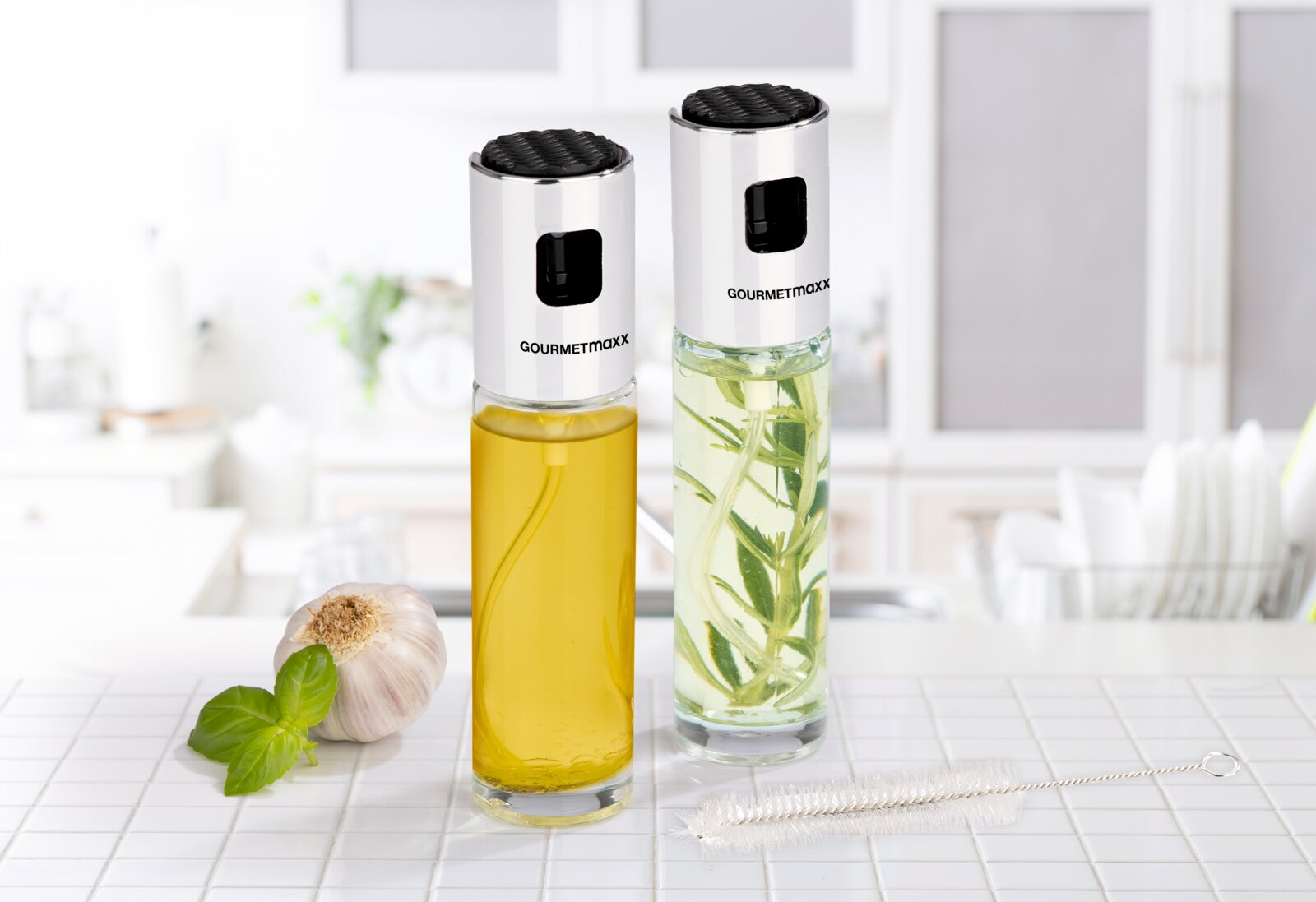 Zwei Gourmetmaxx Öl- und Essigsprüher aus Glas mit silberfarbenen Aufsätzen und schwarzen Druckknöpfen, frontal aus leichter Obersicht neben Knoblauch, Basilikum und Reinigungsbürste präsentiert