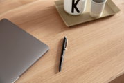 Nahaufnahme eines Schreibtisches aus Holz mit einem Laptop, einem schwarzen Kugelschreiber und einer Tasse auf einem Tablett. Perspektive von oben.