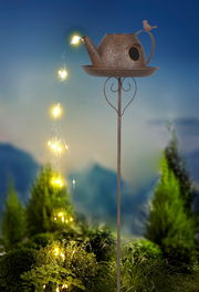 Solarleuchte in Form einer Gießkanne mit Lichterkette, die von oben nach unten fließt, auf einem Stab montiert, vor einem Hintergrund mit Pflanzen, seitliche Perspektive.