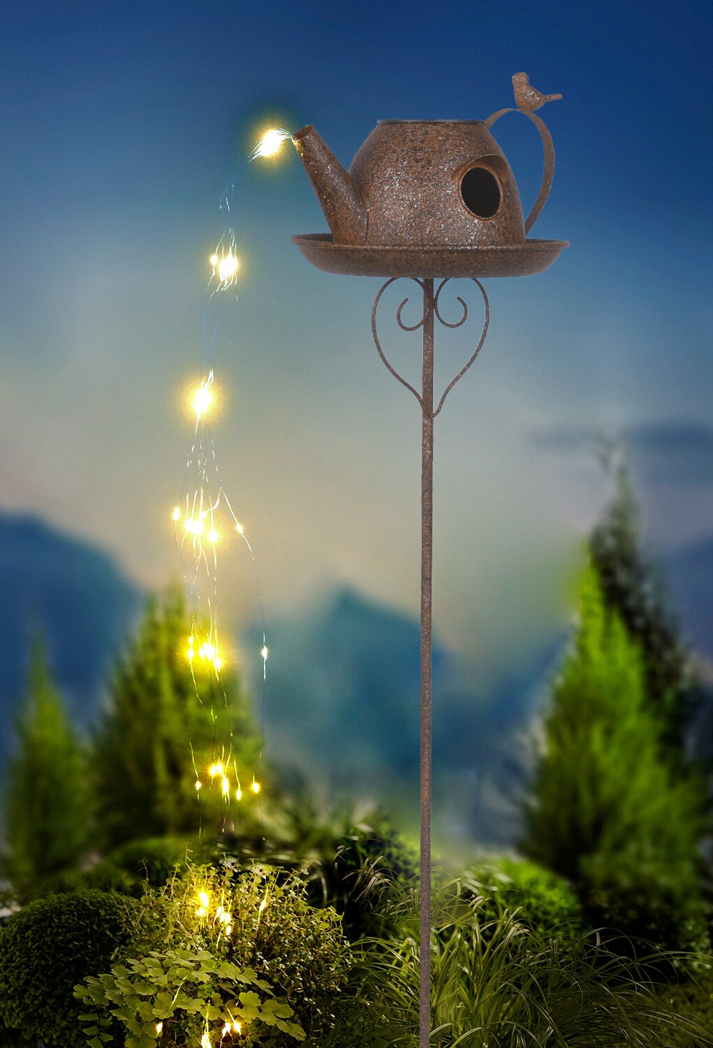 Ambia Garden Solarleuchte SOLARLEUCHTE Solarleuchte in Form einer Gießkanne mit Lichterkette, die von oben nach unten fließt, auf einem Stab montiert, vor einem Hintergrund mit Pflanzen, seitliche Perspektive.