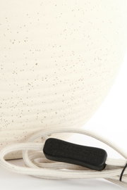 Nahaufnahme eines cremefarbenen Lampenfußes mit feiner Sprenkelung und einem weißen Kabel mit schwarzem Schalter, seitliche Perspektive.