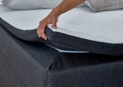 Nahaufnahme eines Boxspringbetts mit einer Hand, die die Matratze anhebt, um die Struktur und den Bezug zu zeigen.