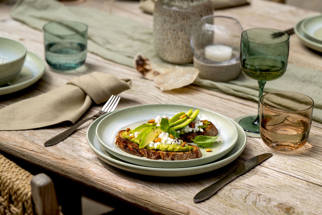 Frühstücksteller mit Avocado-Toast, von schräg oben fotografiert, auf einem gedeckten Tisch mit grünem Geschirr und Gläsern.