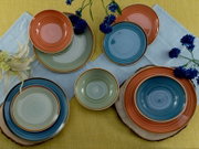 Buntes Keramik-Geschirrset mit Tellern und Schalen in Blau, Grün und Terrakotta auf gelbem Tischtuch mit hellblauem Läufer und blauen Blumen, Draufsicht von oben