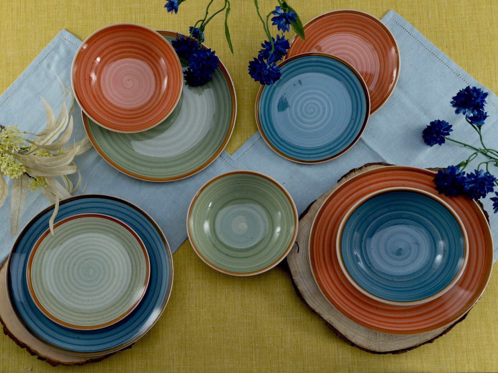 Buntes Keramik-Geschirrset mit Tellern und Schalen in Blau, Grün und Terrakotta auf gelbem Tischtuch mit hellblauem Läufer und blauen Blumen, Draufsicht von oben