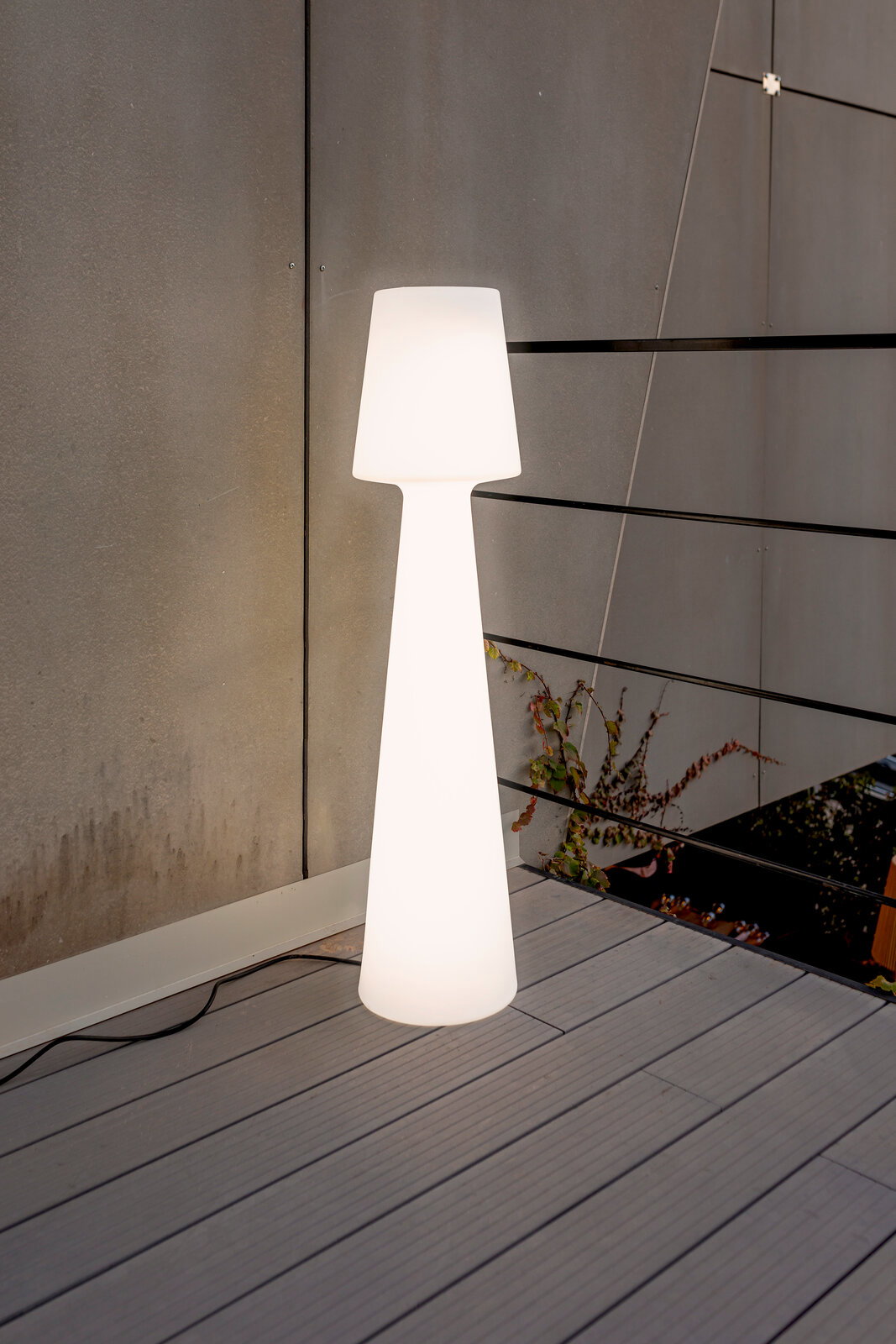 Leuchtende weiße Outdoor-Stehleuchte in schlanker, konischer Form auf einer Terrasse vor grauer Wand und Geländer, frontale Perspektive.