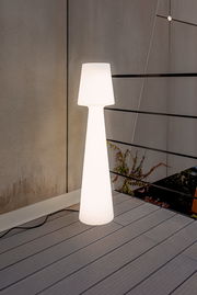 Leuchtende weiße Outdoor-Stehleuchte in schlanker, konischer Form auf einer Terrasse vor grauer Wand und Geländer, frontale Perspektive.