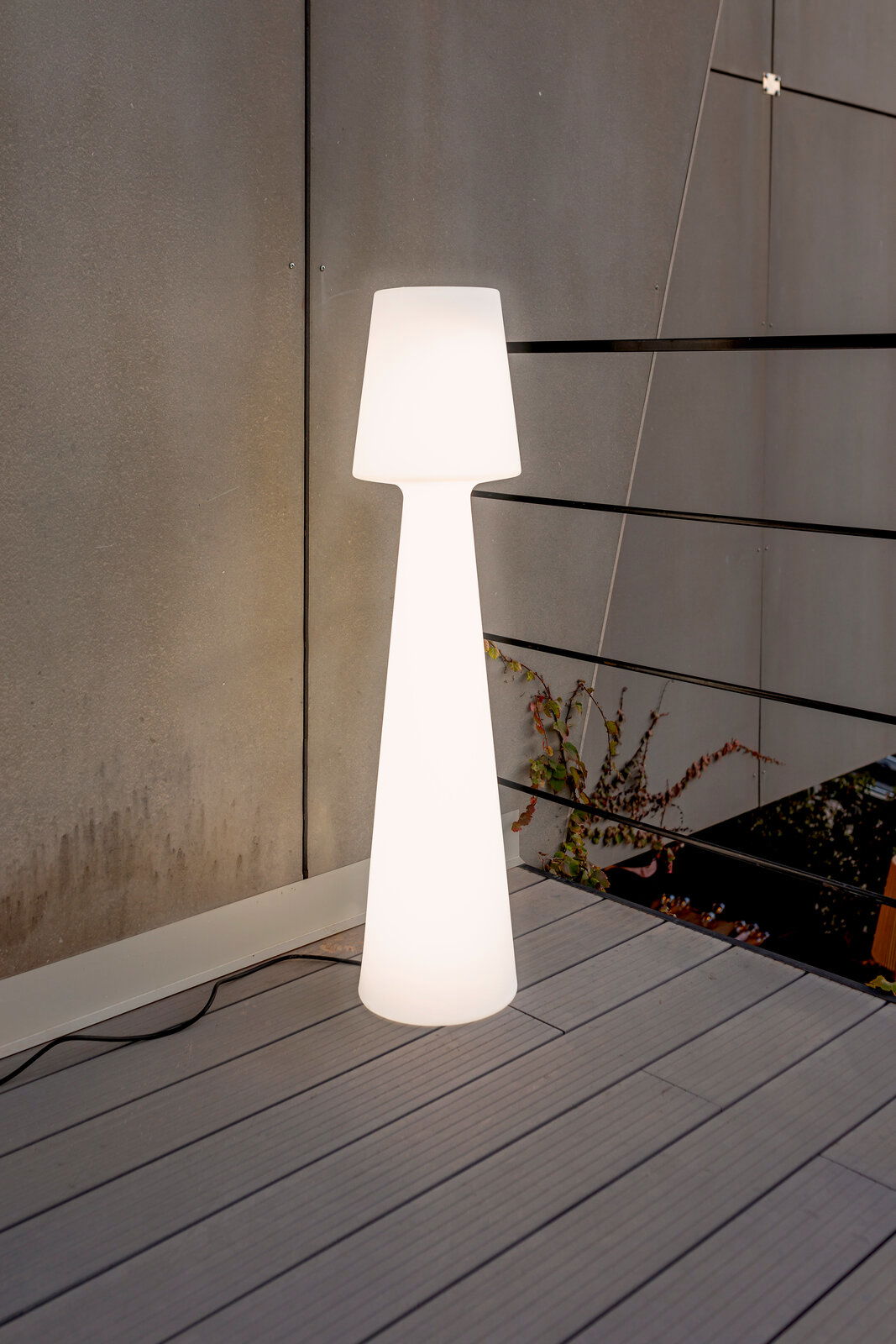 Leuchtende weiße Outdoor-Stehleuchte in schlanker, konischer Form auf einer Terrasse vor grauer Wand und Geländer, frontale Perspektive.