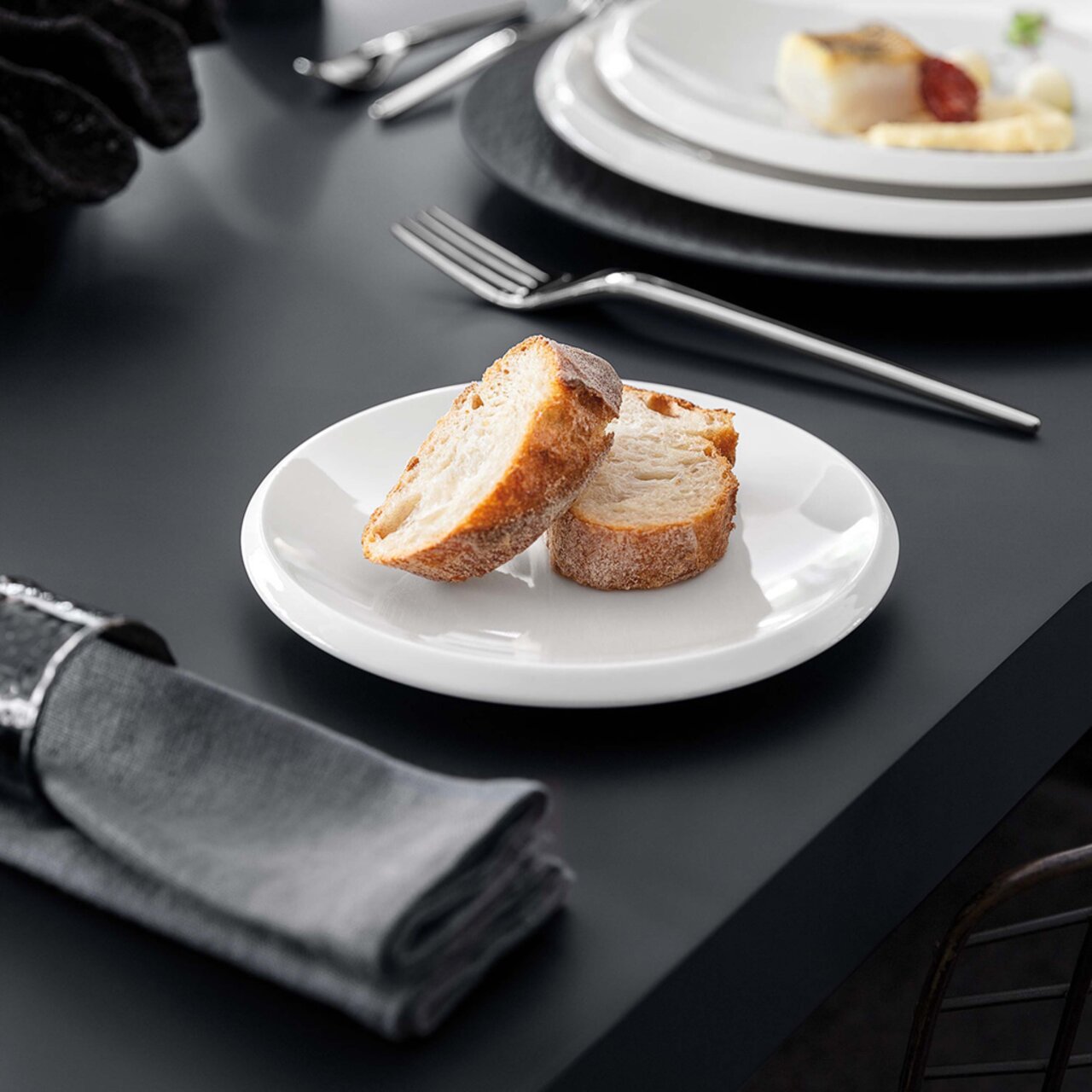 Weißer Brotteller NewMoon 16cm mit zwei Brotscheiben, seitliche Perspektive auf einem gedeckten Tisch.