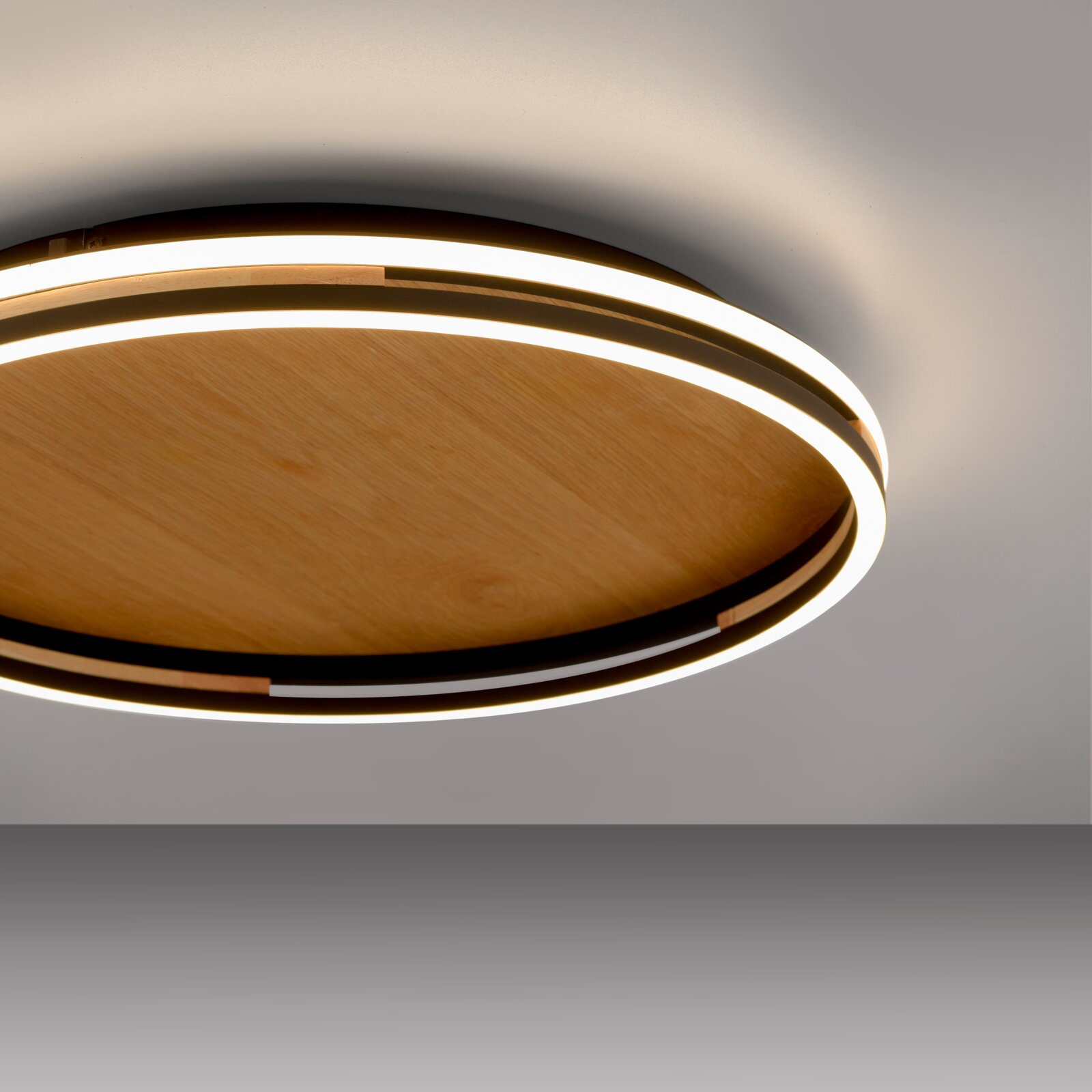 Moderne runde LED-Deckenleuchte mit Holzoptik und umlaufendem Lichtband, fotografiert aus schräger Untersicht (Blick von unten nach oben) vor hellem Hintergrund