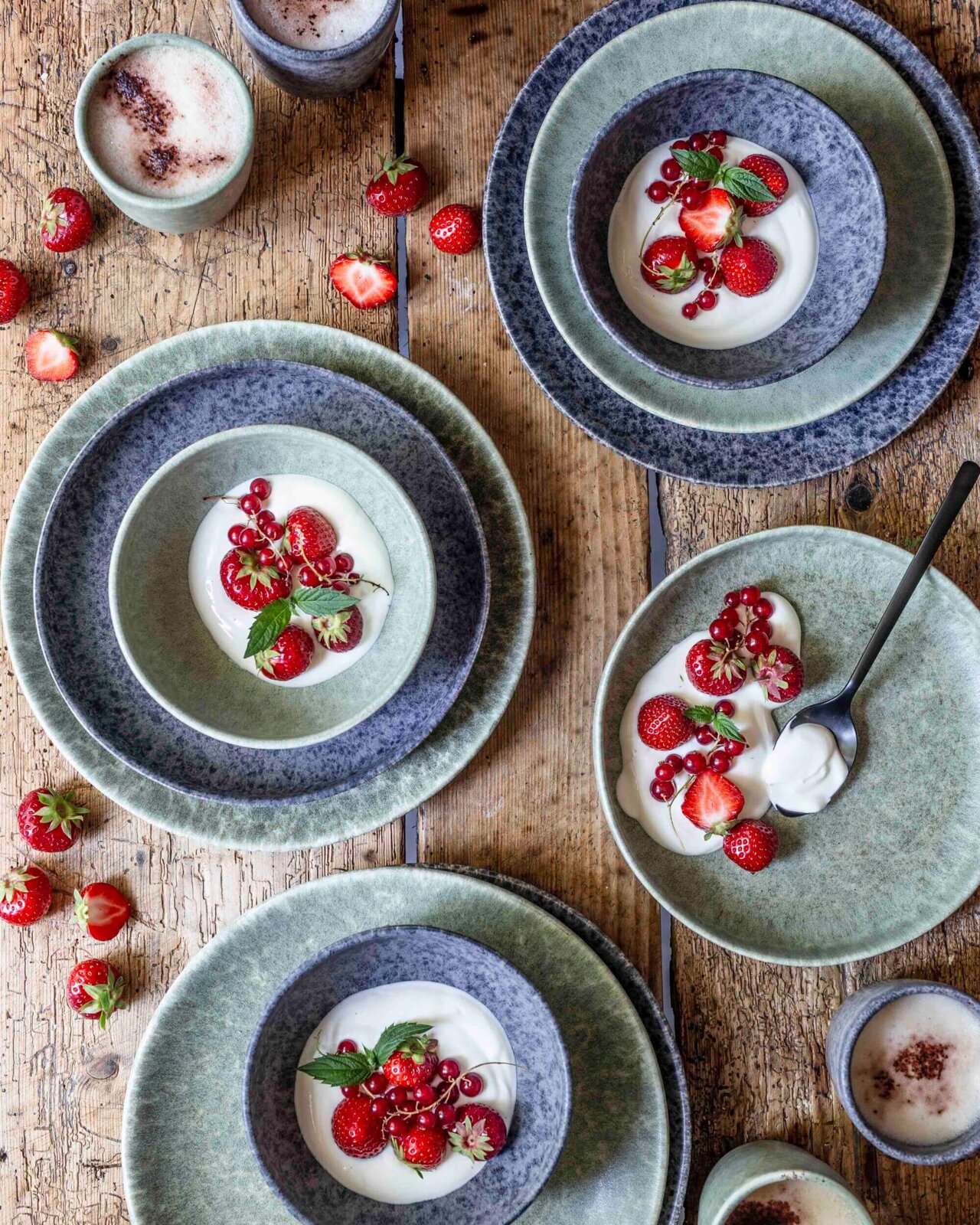 Draufsicht auf rustikal gedeckten Holztisch mit grün-blauem Keramikgeschirr: mehrere Teller mit weißem Dessert, garniert mit Erdbeeren, roten Johannisbeeren und Minze, daneben Tassen mit schaumigem Getränk.