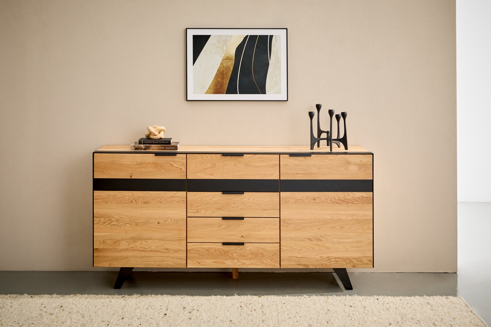 Modern houten dressoir met zwarte accenten, gefotografeerd vanaf de voorkant, versierd met boeken en kandelaars, tegen een beige muur met daarboven een abstract kunstwerk.