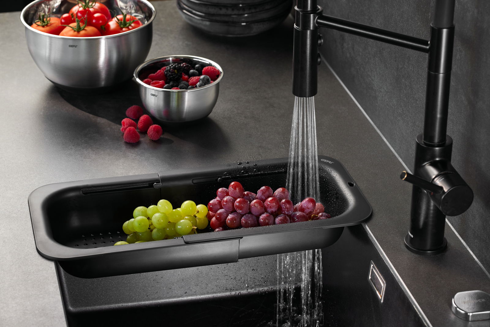 GEFU Spülbeckensieb DRENARE Spülbeckensieb DRENARE mit frischen Trauben, von oben fotografiert, während Wasser aus einem schwarzen Wasserhahn fließt.
