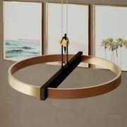 Moderne ronde hanglamp met goudkleurige ring en zwarte dwarsbalk, van schuin onderen gezien tegen een wand met strandposters.