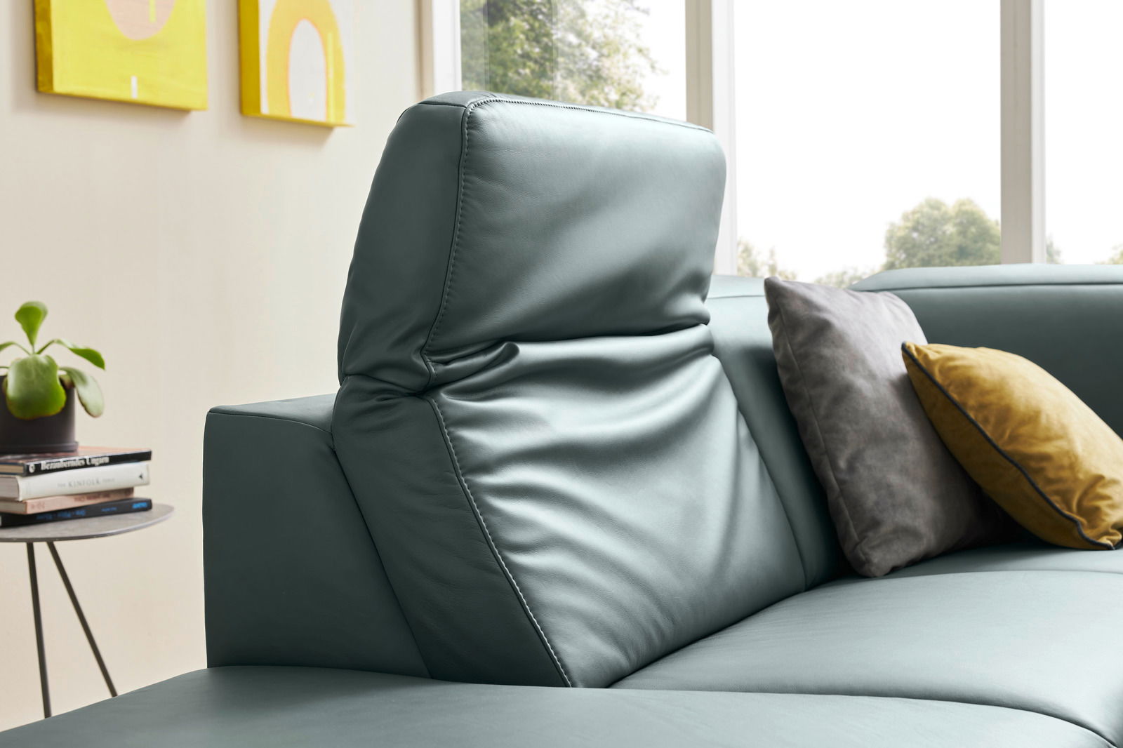 Nahaufnahme einer modernen Eckgarnitur mit verstellbarer Kopfstütze in grünem Leder, dekoriert mit grauen und gelben Kissen, seitliche Perspektive.
