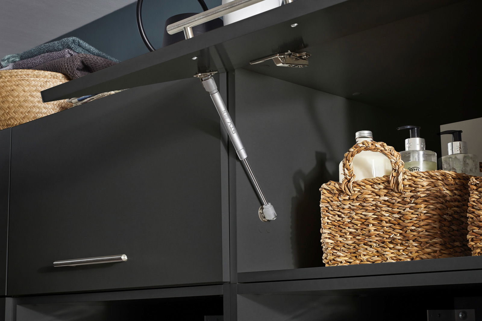 Nahaufnahme eines geöffneten, breiten Aufsatzschranks mit dunkler Oberfläche, gefüllt mit geflochtenen Körben und Pflegeprodukten, von unten betrachtet.