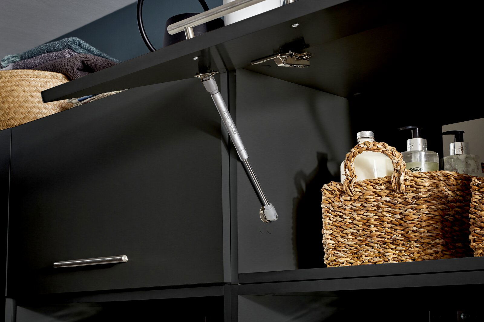 Nahaufnahme eines geöffneten, breiten Aufsatzschranks mit dunkler Oberfläche, gefüllt mit geflochtenen Körben und Pflegeprodukten, von unten betrachtet.