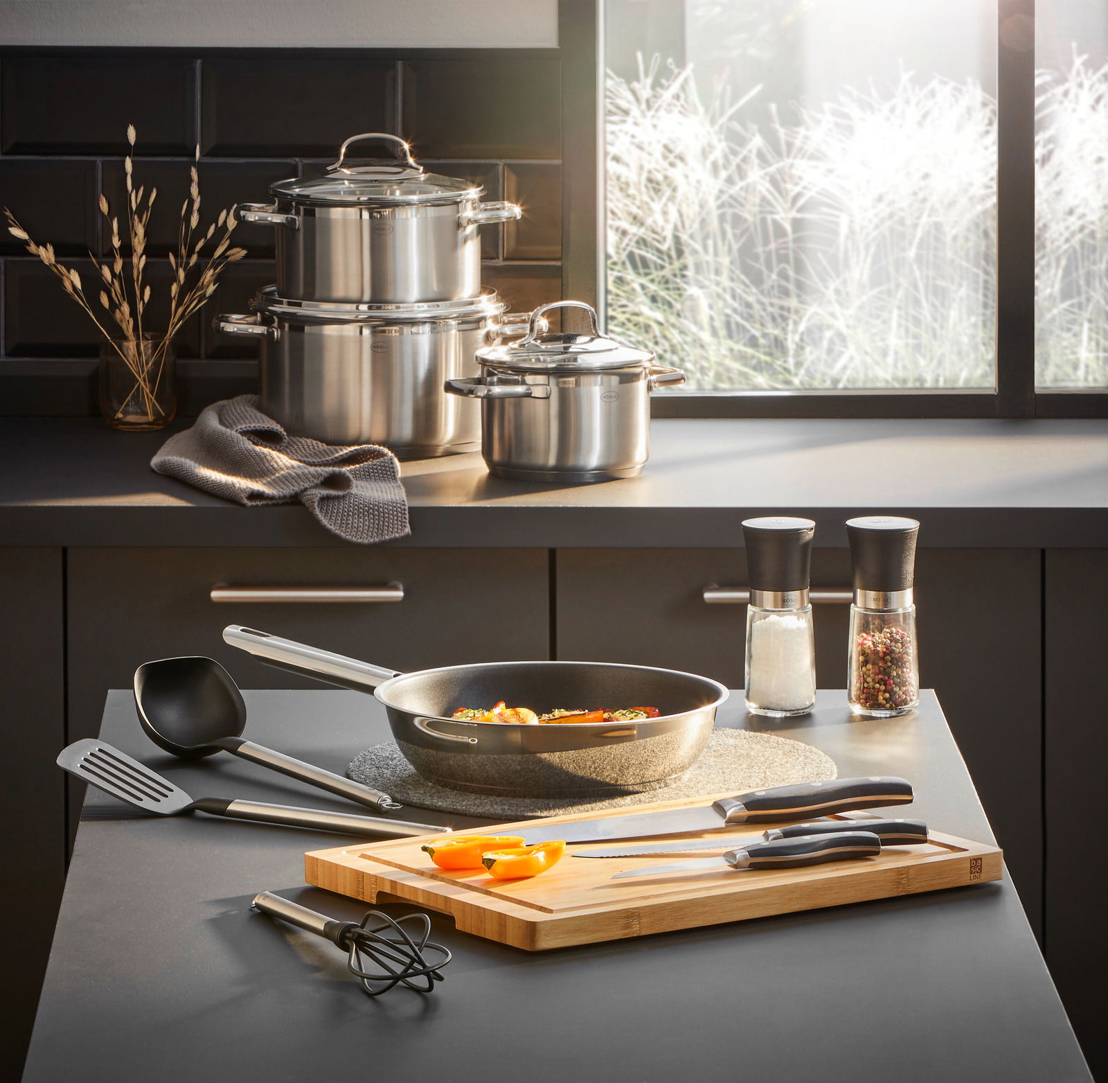 Küchenansicht mit Gourmet-Set Elegance 13-tlg., bestehend aus Töpfen, Pfanne, Küchenutensilien und Schneidebrett, aus der Perspektive einer modernen Küche.