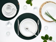 Draufsicht auf ein modern gedecktes Tisch-Arrangement mit weißen Tellern und Tasse, Besteck und Wassergläsern auf dunkelgrünen Platzsets, ergänzt durch Kerzen, grüne Blätter und eine Holzplatte mit Schüssel auf weißem Hintergrund.