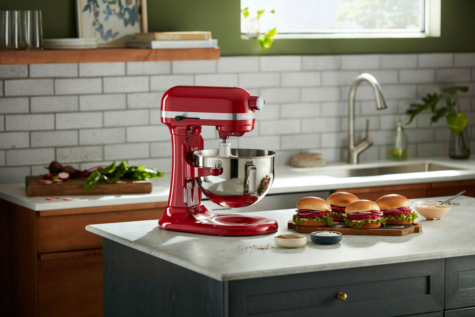 KitchenAid Küchenmaschine HEAVY DUTY 5KSM55SXXEER Rote Küchenmaschine Heavy Duty 5,2L auf einer Küchenarbeitsplatte, seitliche Perspektive, mit Zutaten und Sandwiches im Hintergrund.