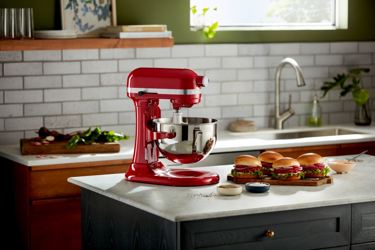 Rote Küchenmaschine Heavy Duty 5,2L auf einer Küchenarbeitsplatte, seitliche Perspektive, mit Zutaten und Sandwiches im Hintergrund.