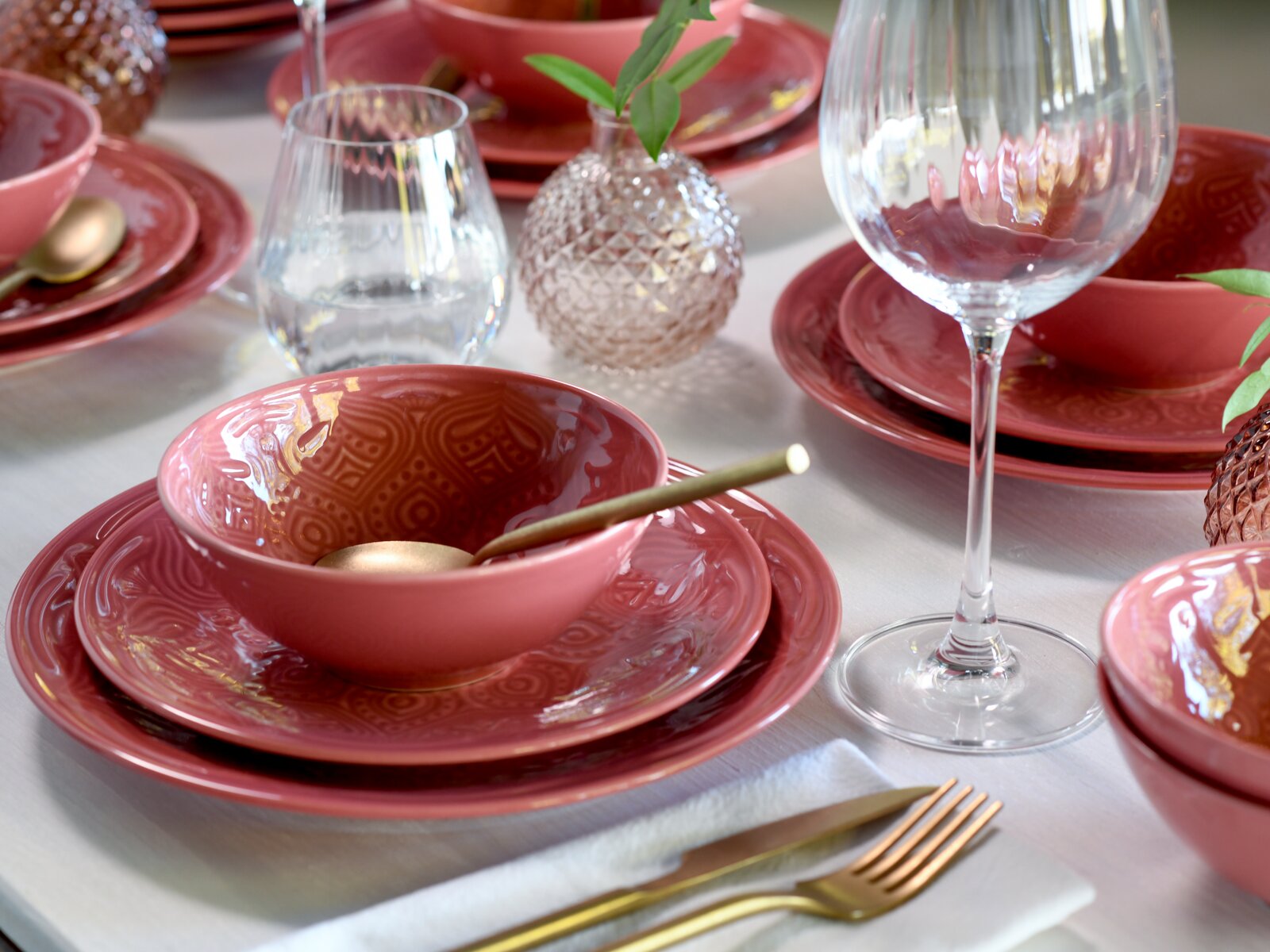 Gedeckter Tisch mit roséfarbenem, geprägtem Keramik-Geschirr (Teller und Schalen), goldfarbenem Besteck und Weingläsern, fotografiert aus leicht schräger Seitenansicht (3/4-Perspektive)