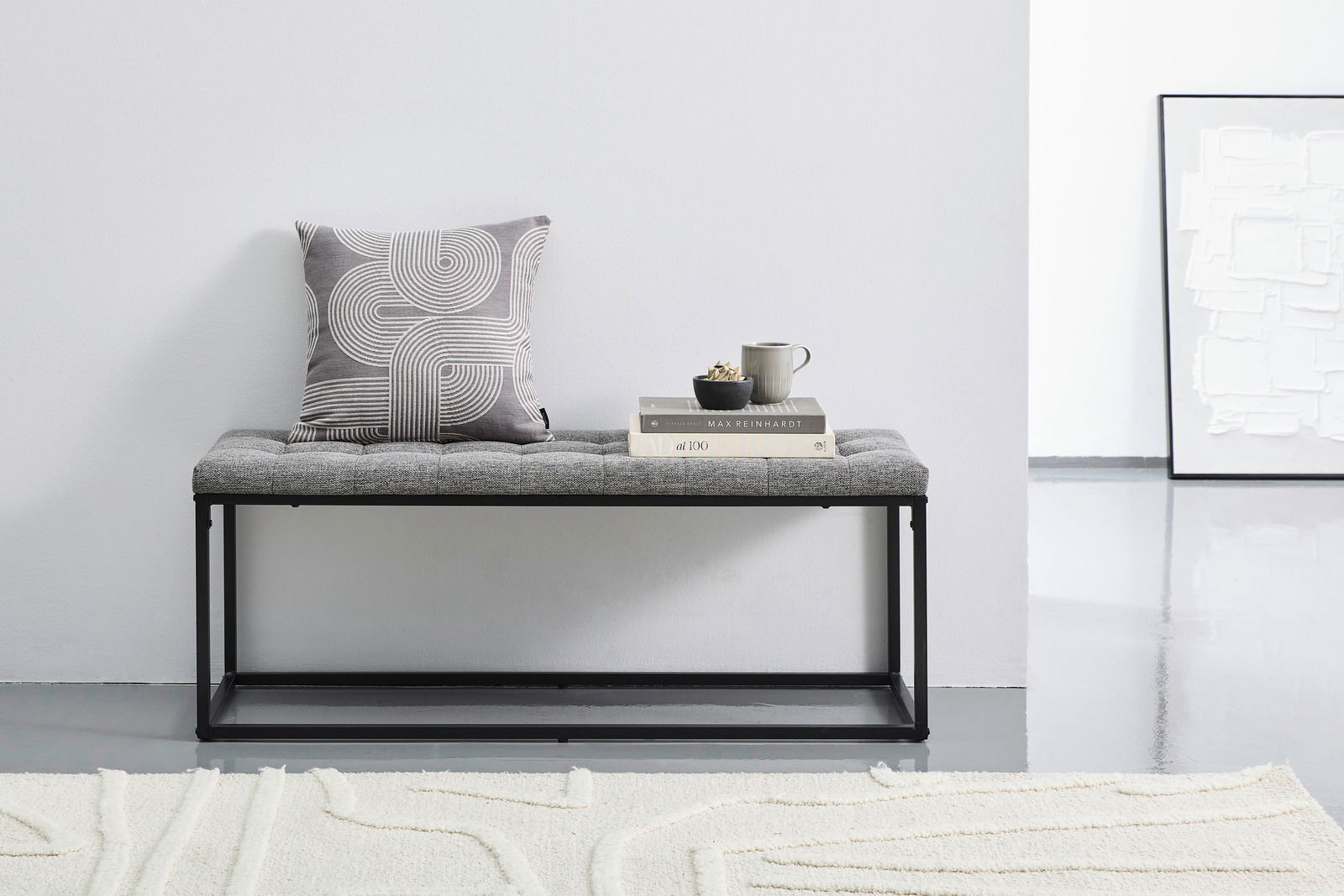 Graue Polsterbank mit schwarzem Metallrahmen, dekoriert mit einem Kissen und Büchern, seitliche Perspektive.