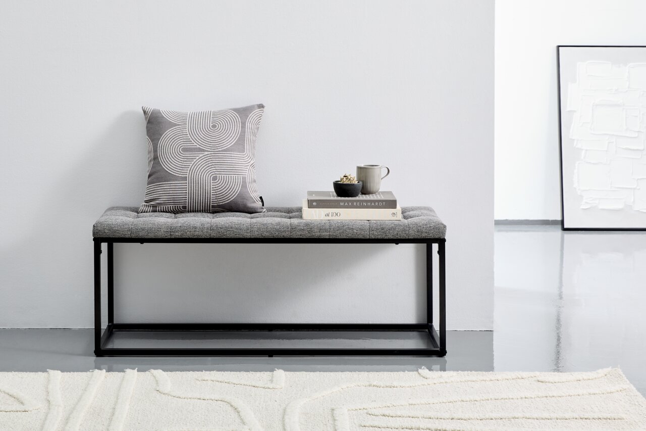 Graue Polsterbank mit schwarzem Metallrahmen, dekoriert mit einem Kissen und Büchern, seitliche Perspektive.