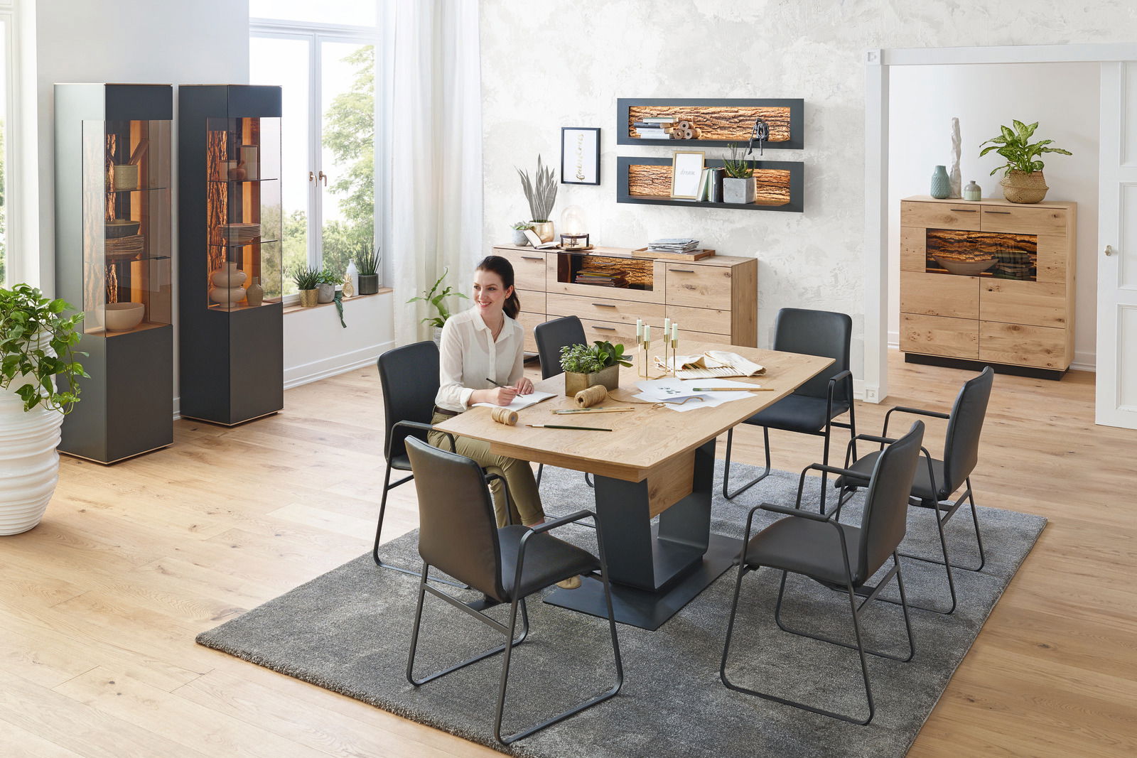 Moderner Essbereich mit Esstisch aus Holz und Metall, umgeben von schwarzen Stühlen. Im Hintergrund sind Vitrinen, ein Sideboard und ein Wandpaneel zu sehen. Die Perspektive zeigt den Raum aus einer leicht erhöhten Position.