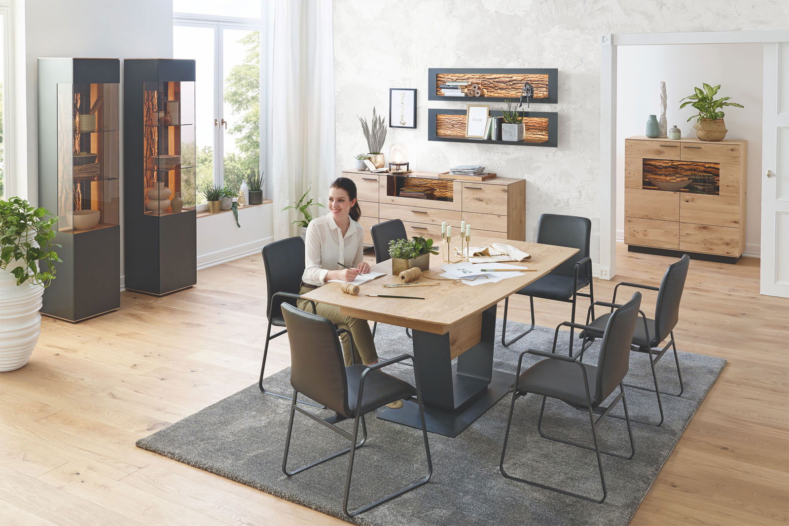 Moderner Essbereich mit Esstisch aus Holz und Metall, umgeben von schwarzen Stühlen. Im Hintergrund sind Vitrinen, ein Sideboard und ein Wandpaneel zu sehen. Die Perspektive zeigt den Raum aus einer leicht erhöhten Position.
